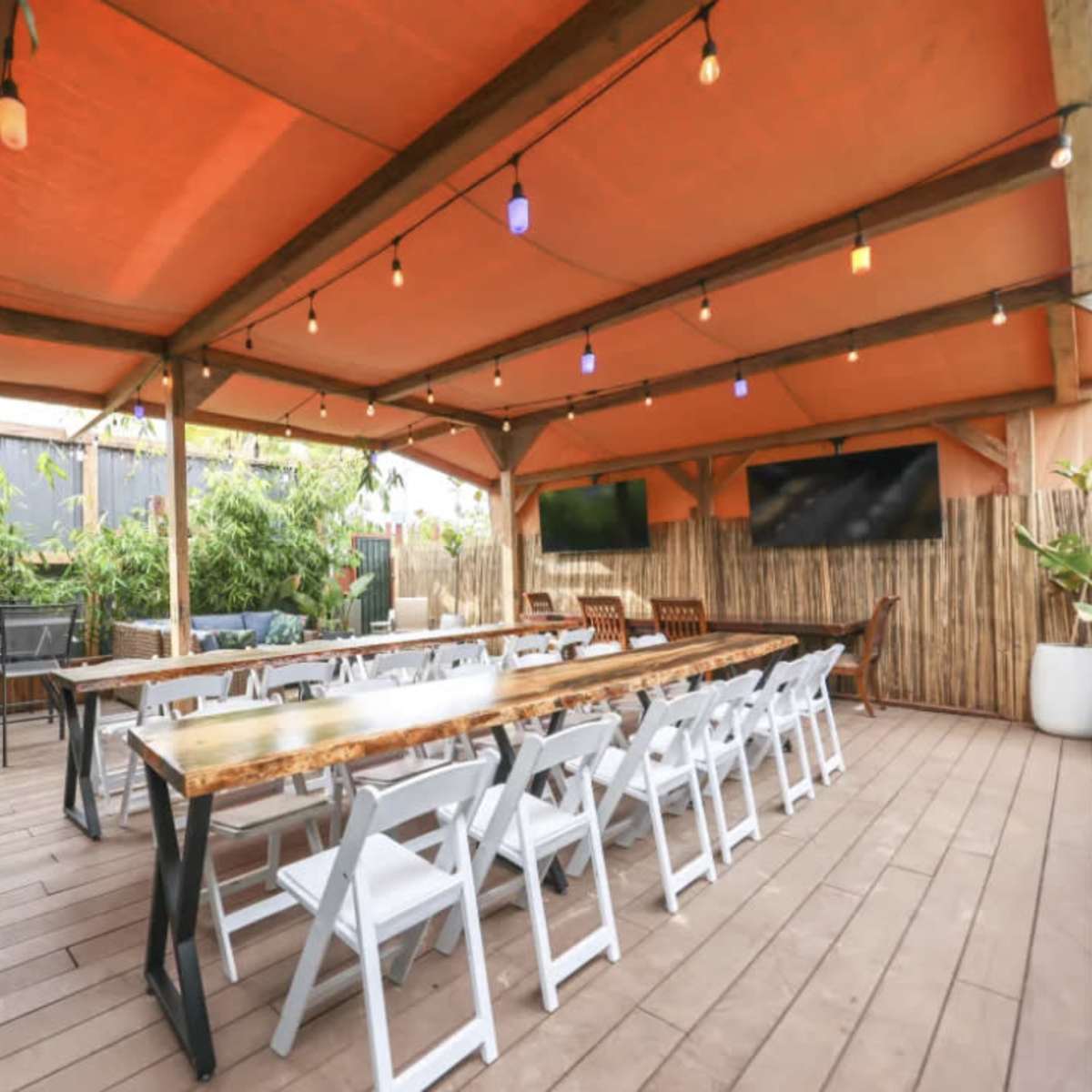 An outdoor dining area with a long wooden table, white folding chairs, string lights, and a grill under a large orange canopy.