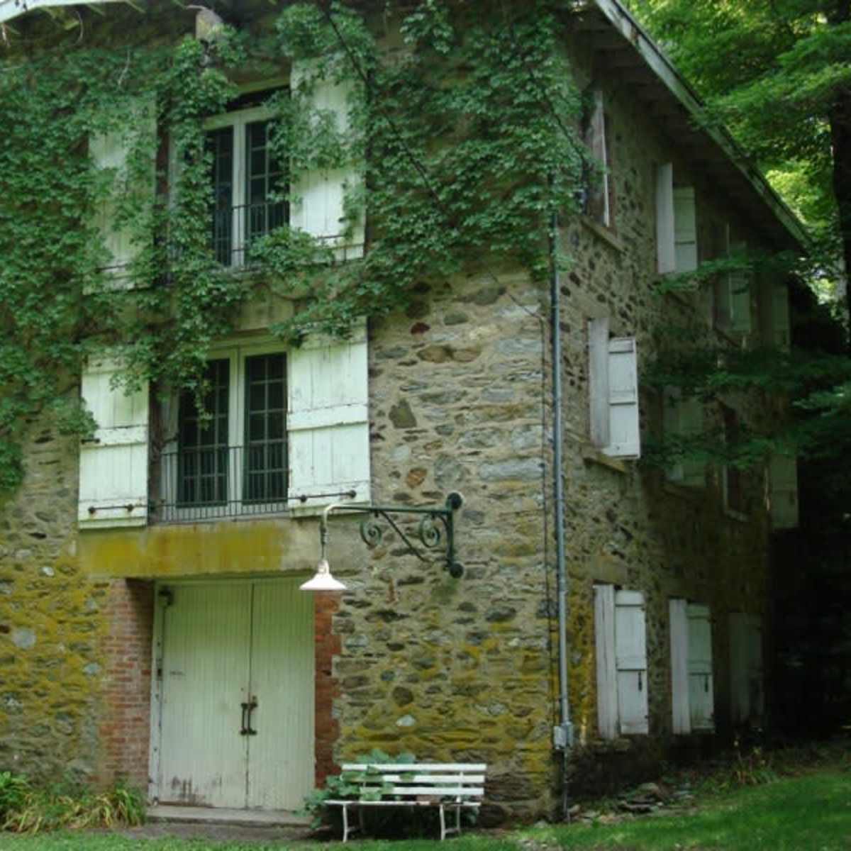 A stone house with green shutters, surrounded by ivy, has a porch light and a white bench in front of it.