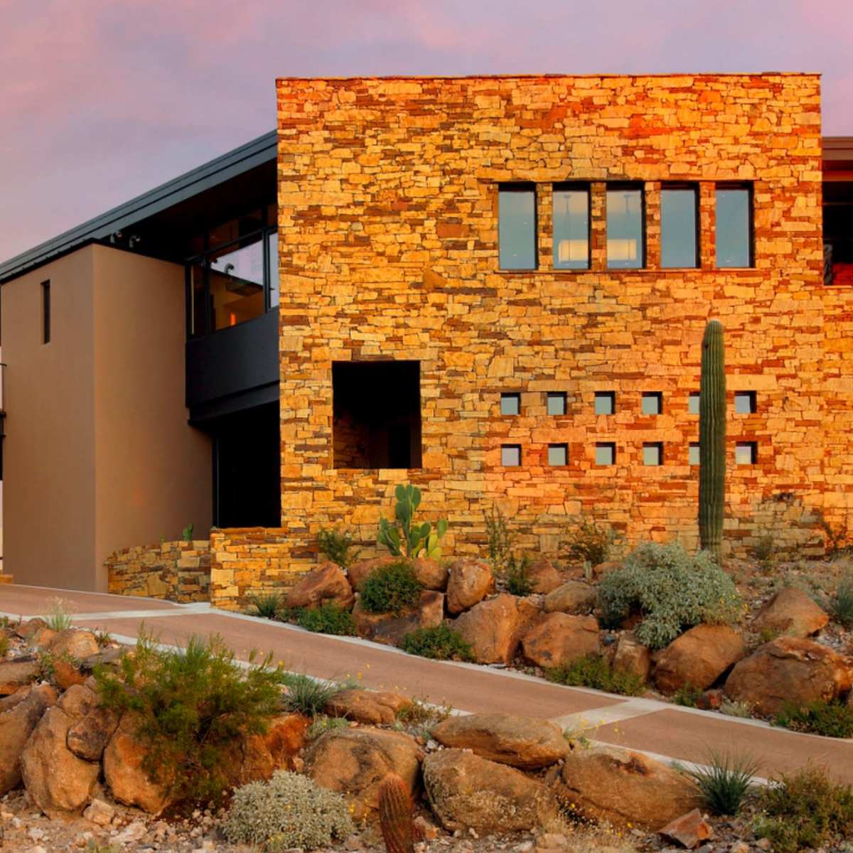 A modern stone house with large windows, set against a desert landscape at sunset.