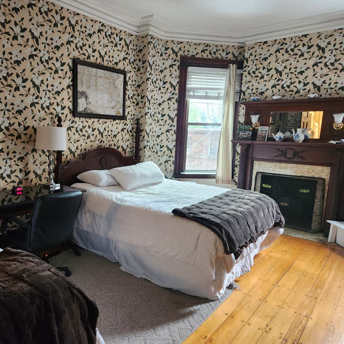 A bedroom with two beds, a chair, a lamp, and a fireplace, all set against floral wallpaper and wooden flooring.