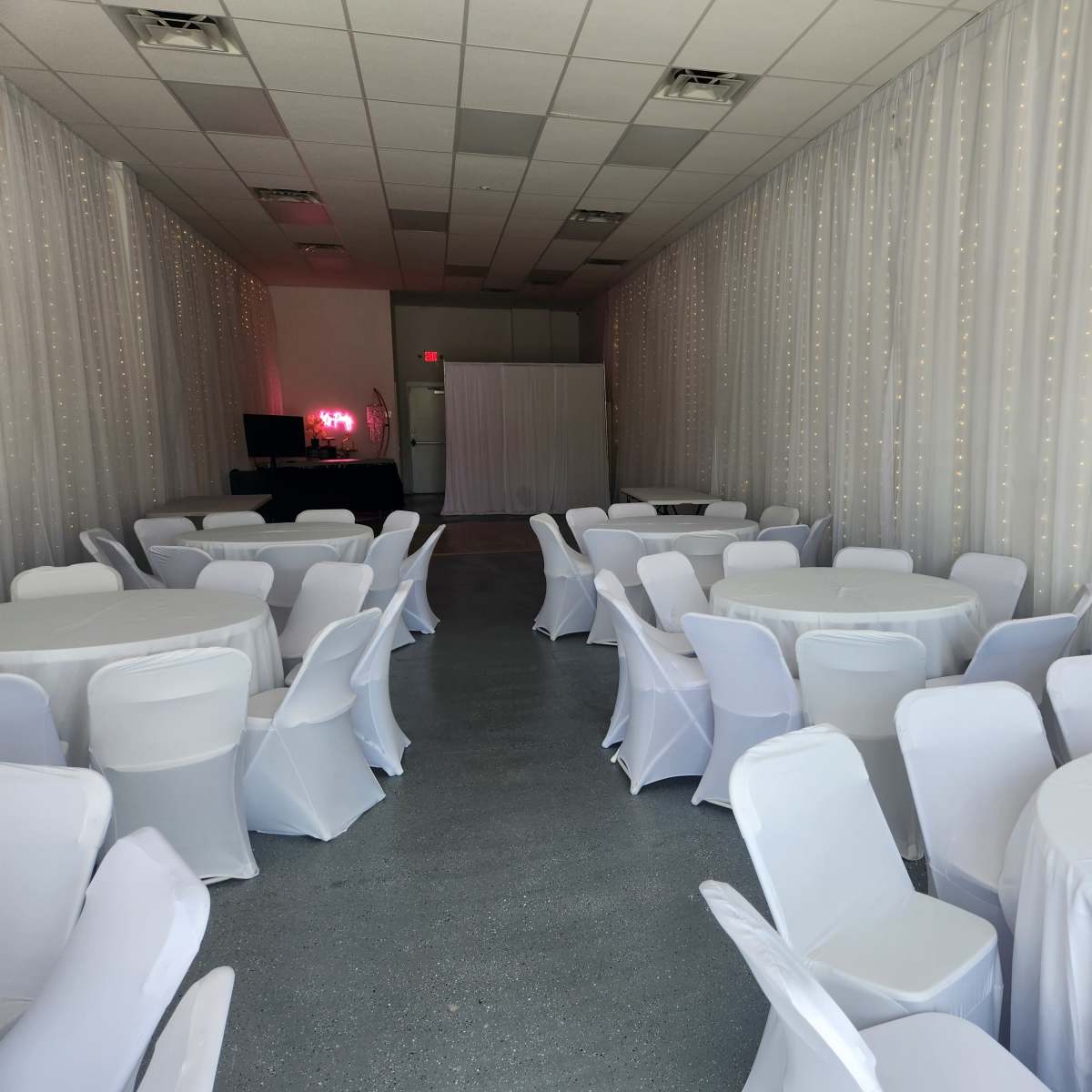 A spacious event room with white tables and chairs arranged neatly under a curtain adorned with small lights.
