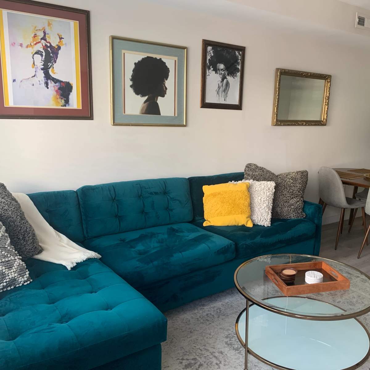 The living area features a teal sofa, a glass coffee table with a wooden tray, framed artwork on the walls, and a dining table with gray chairs in the background.