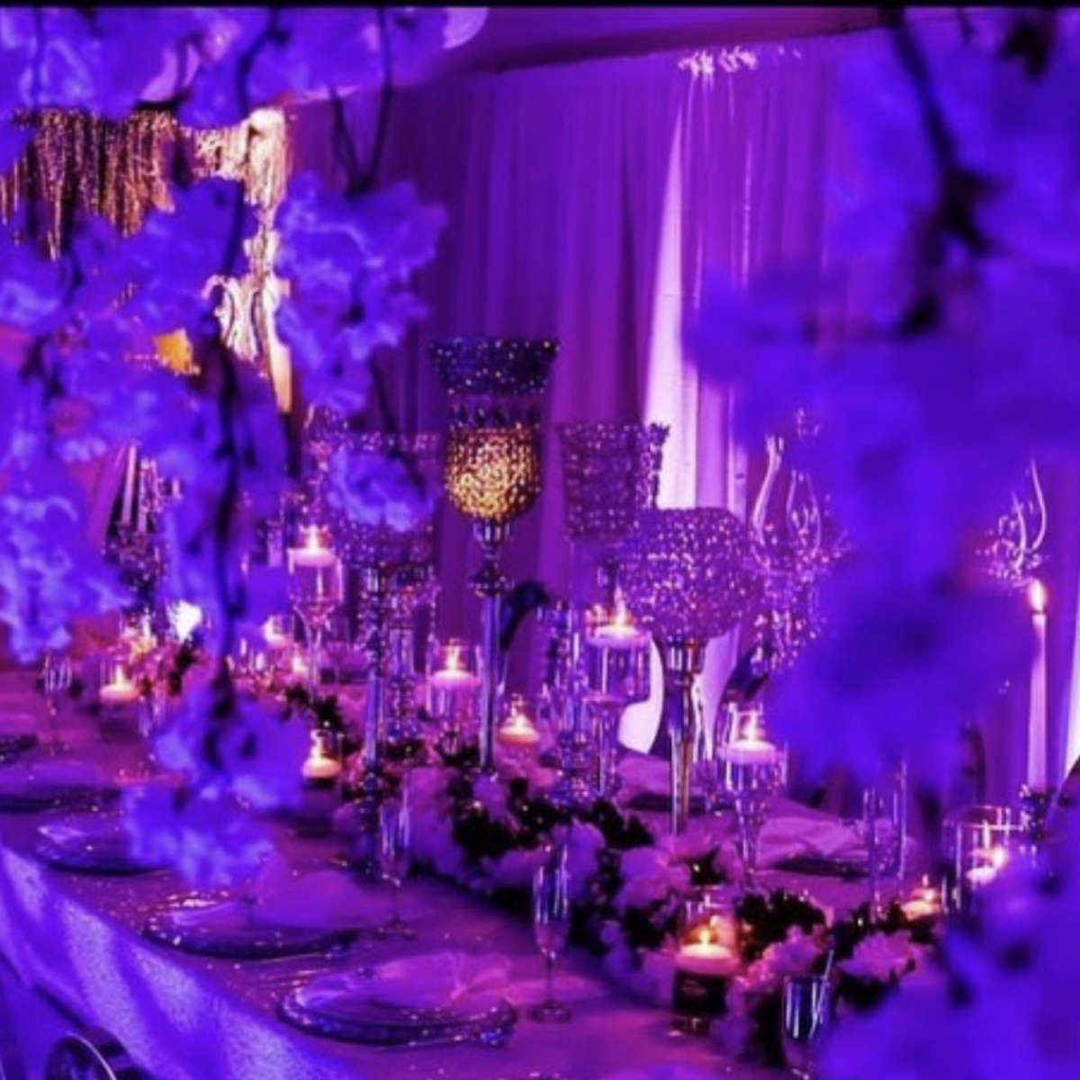 A banquet table is elegantly set with candles and floral arrangements, surrounded by purple-lit decor.