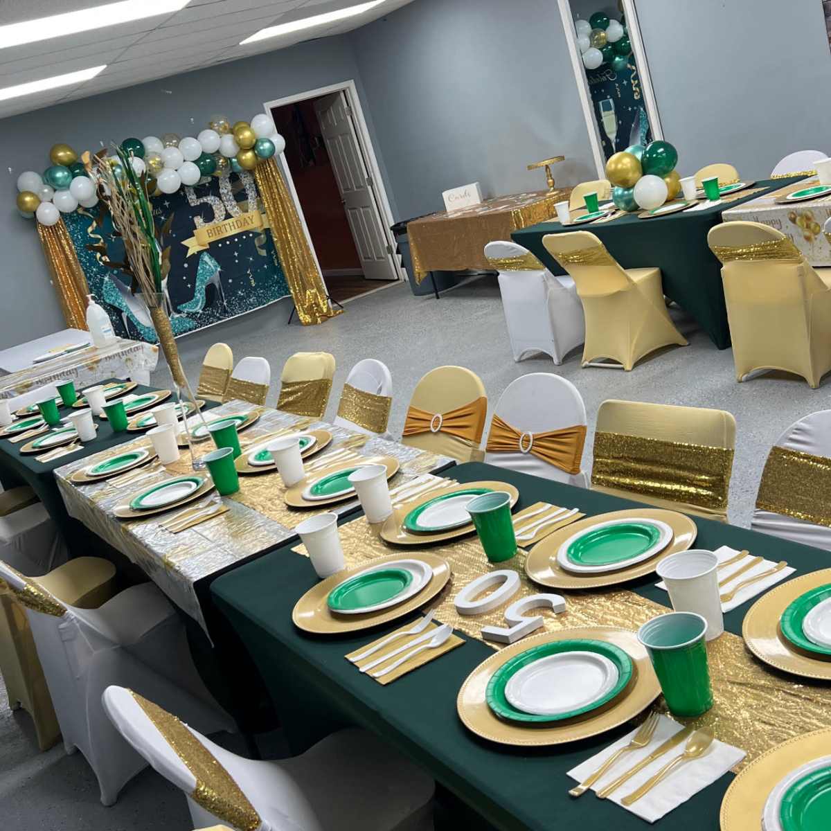 A banquet hall set up for an event, featuring tables decorated with green and gold tableware, white and gold chairs, and a backdrop of balloons.
