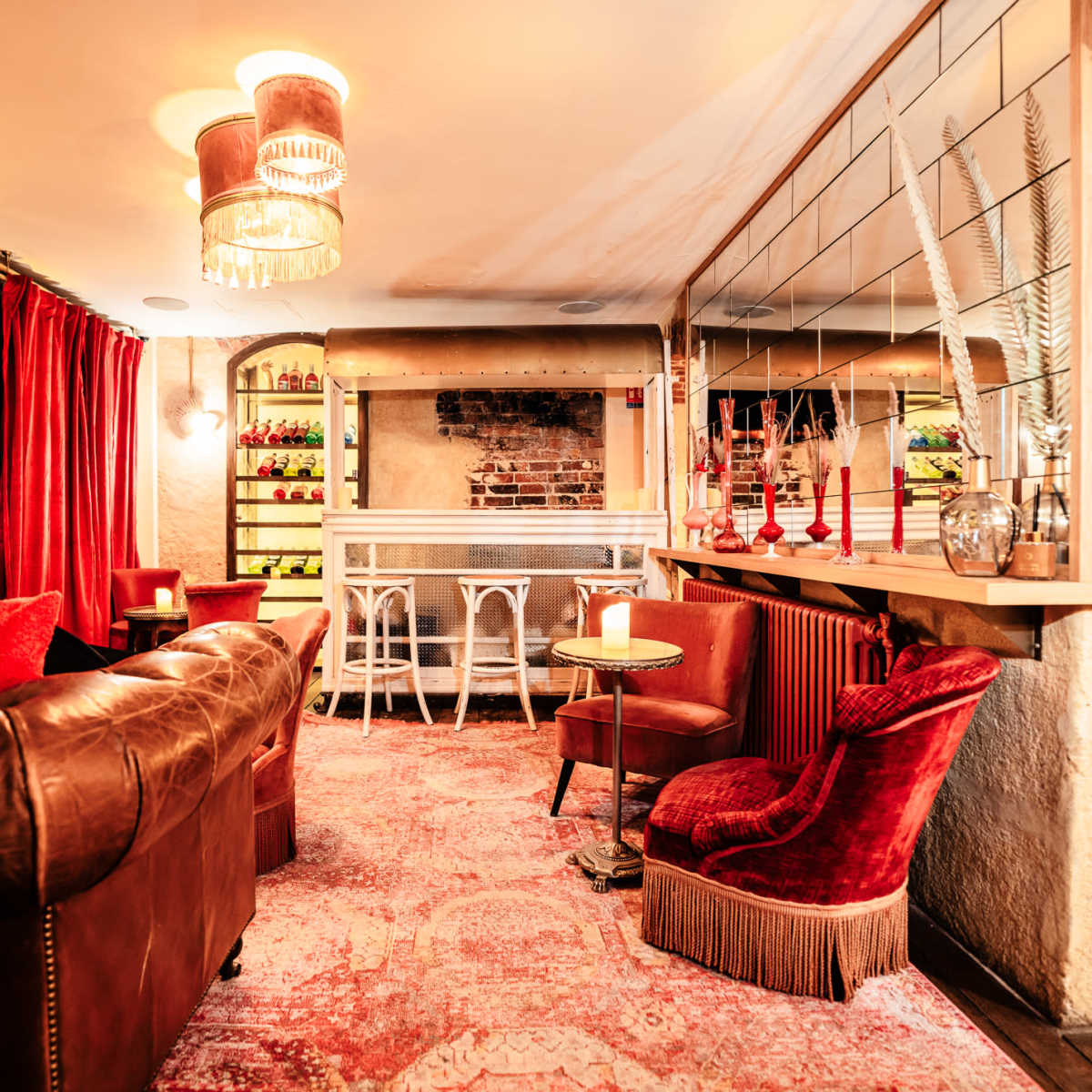 A cozy lounge area featuring a brown leather sofa, a plush red chair, and a small round table, all set against a backdrop of warm lighting and exposed brick walls.