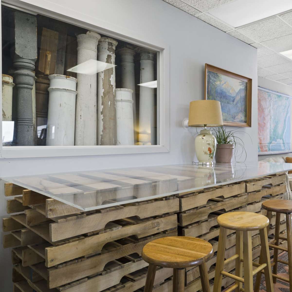 The scene shows a modern interior space featuring a wooden pallet bar with four stools, a cozy armchair, and large windows with natural light.
