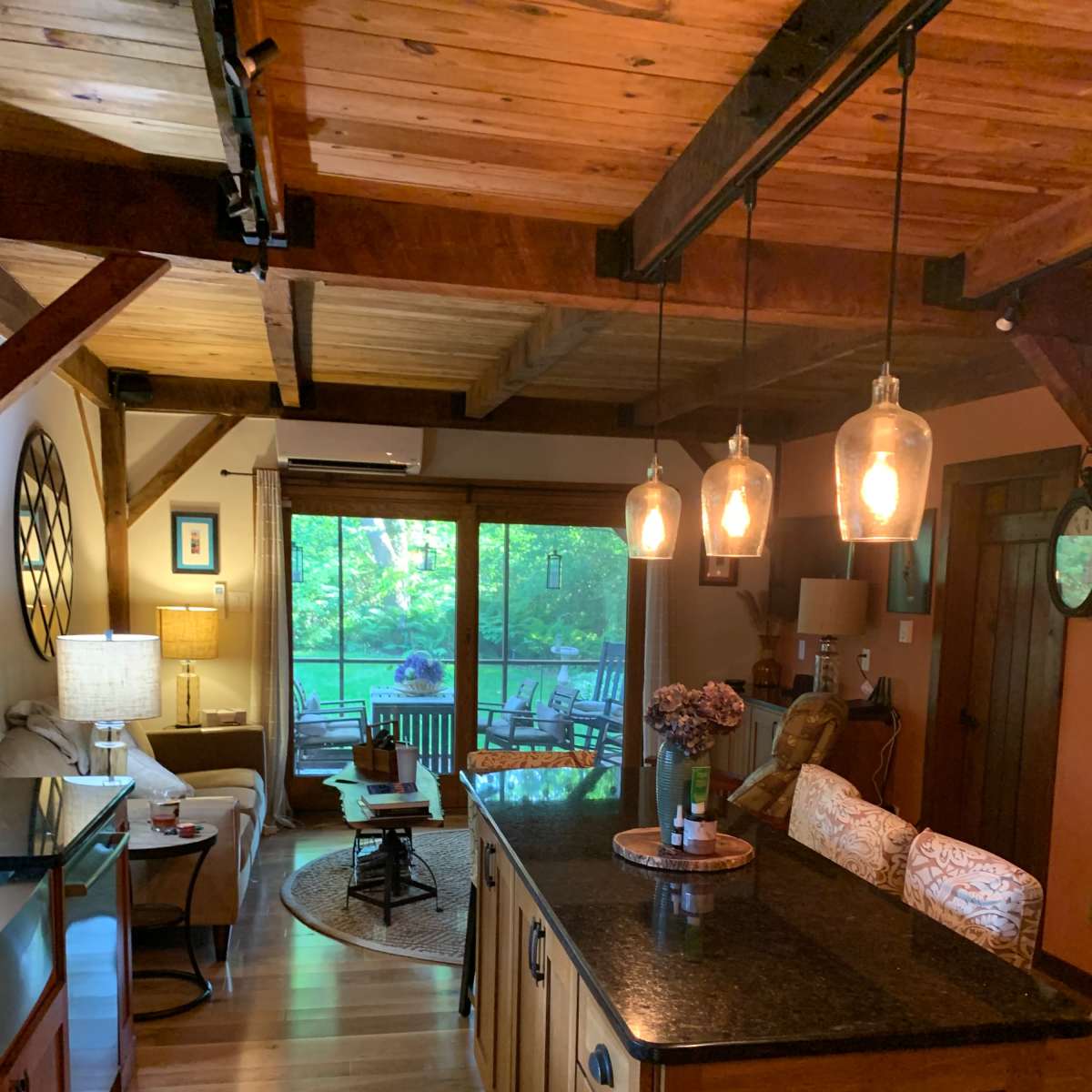 A warm, inviting kitchen opening into a living area with wooden beams and large windows overlooking a green outdoor space.