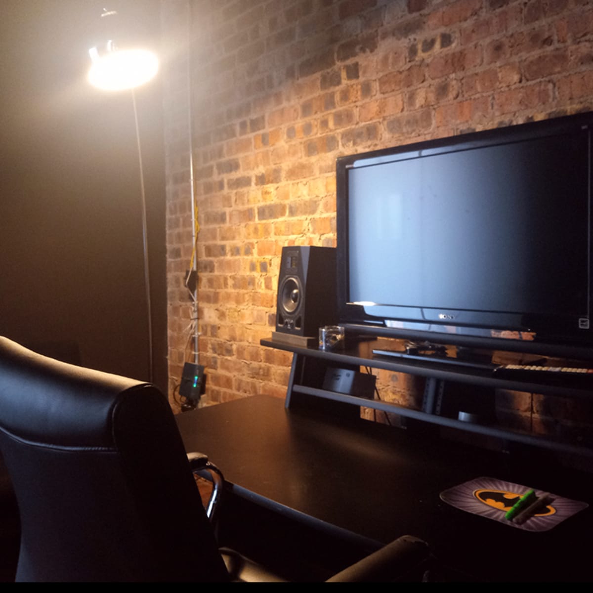 A dark room features a black desk with a computer monitor, speakers, and a swivel chair against a brick wall, illuminated by a hanging light.