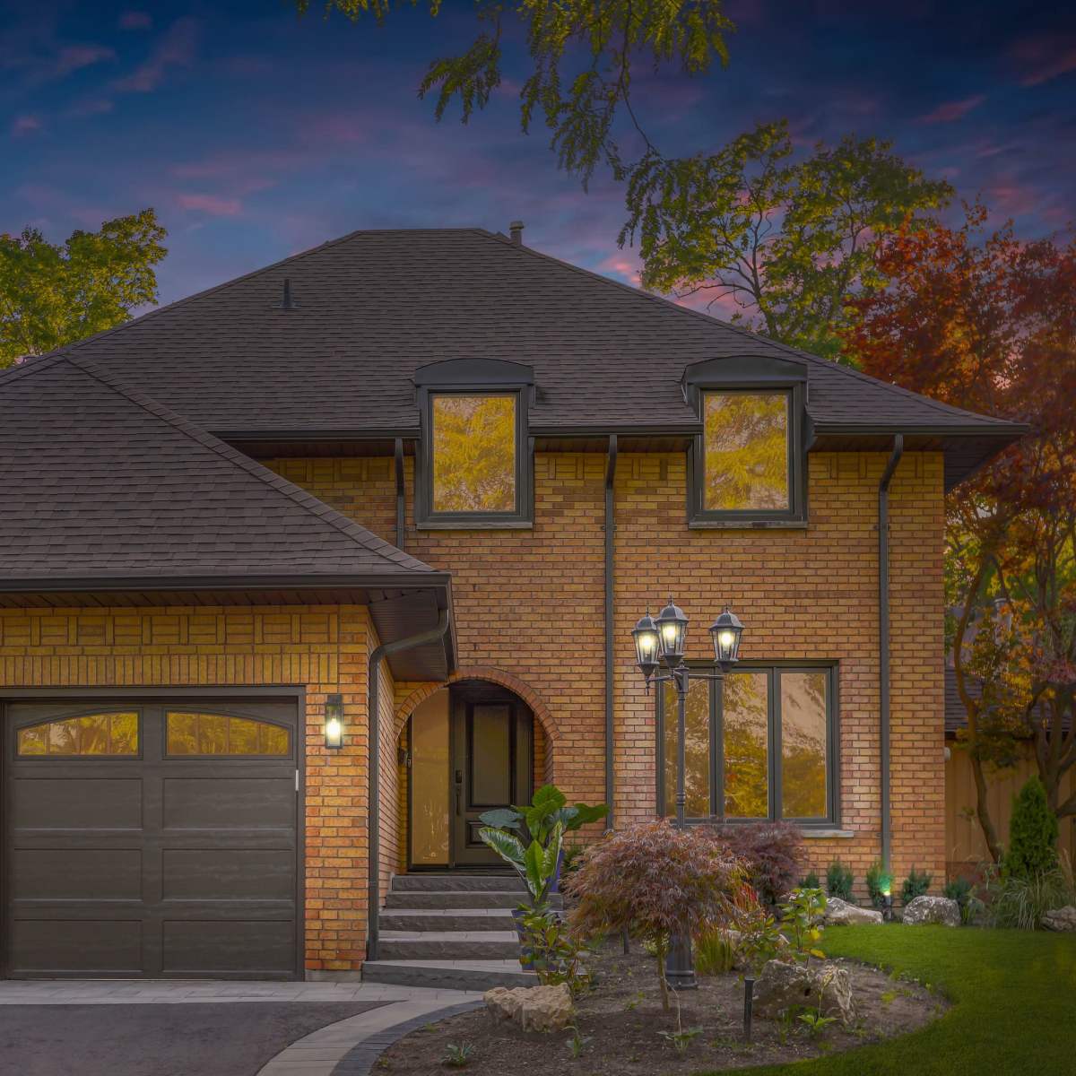 A large brick house with a steep roof, two garage doors, and landscaped greenery in front.