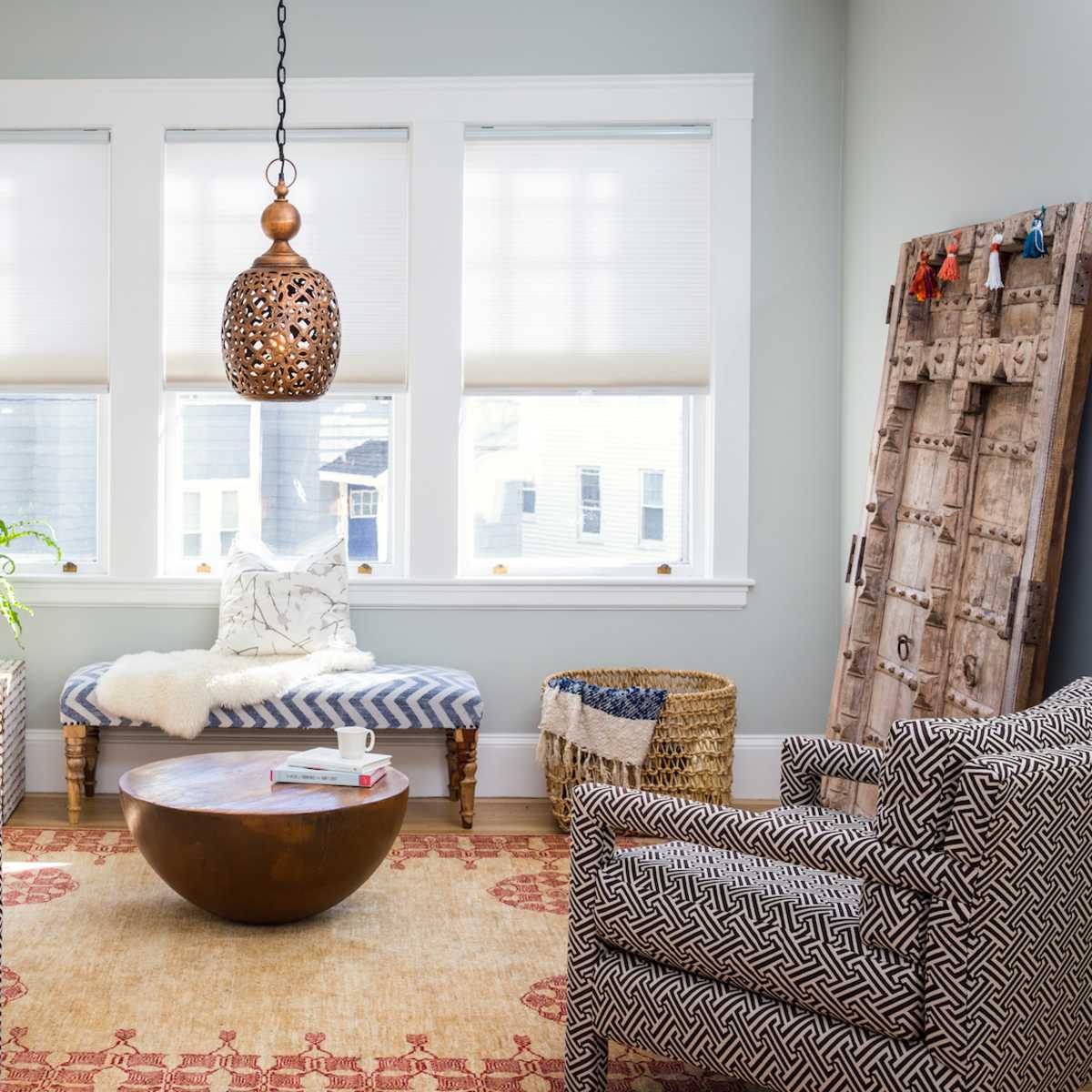A bright living room with two patterned chairs, a round coffee table, and a pendant light, featuring a plant and a woven basket on a patterned rug.