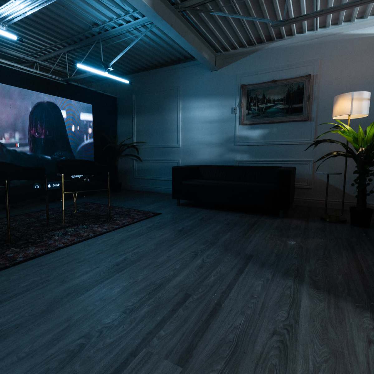 A spacious room with a dark rug, a black sofa, a potted plant, and a large screen displaying an image on one wall.