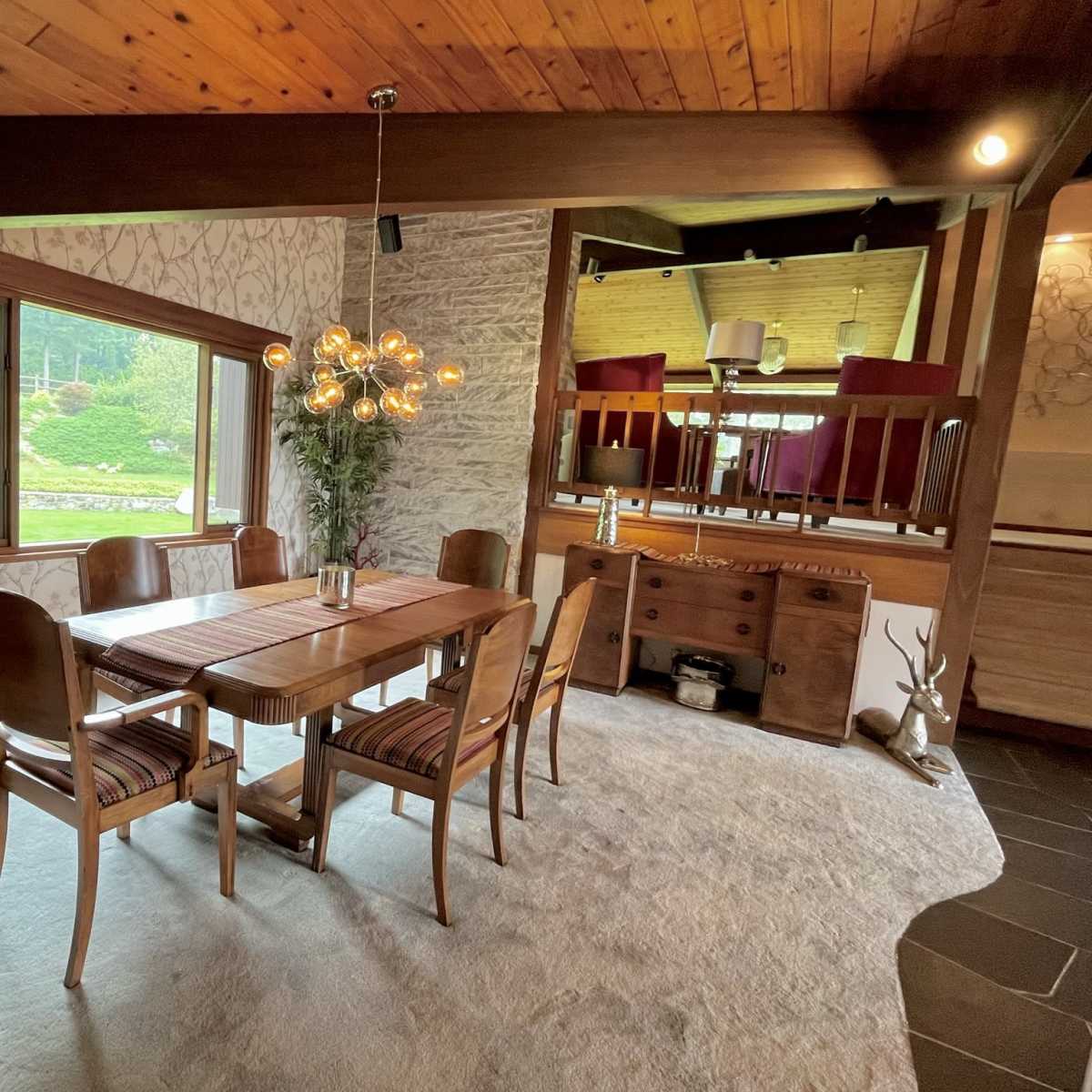 A dining area features a wooden table surrounded by chairs, with a light fixture above and a view of greenery outside through a large window.