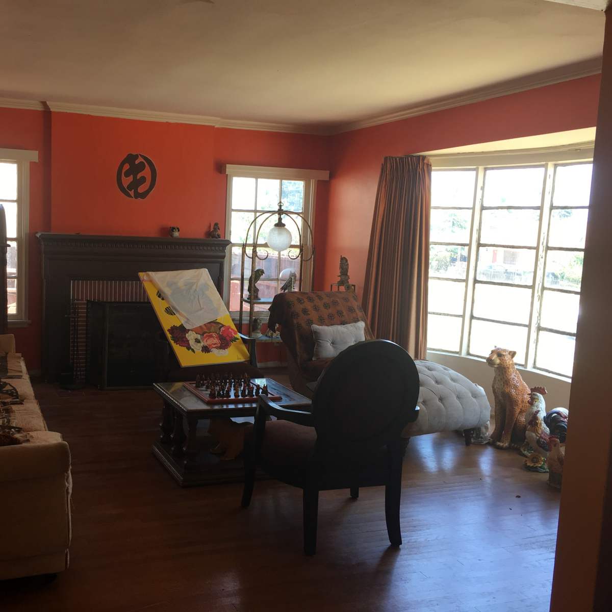 The room features a sofa, a fireplace, a large window, and an easel with a painting, complemented by various furniture pieces.