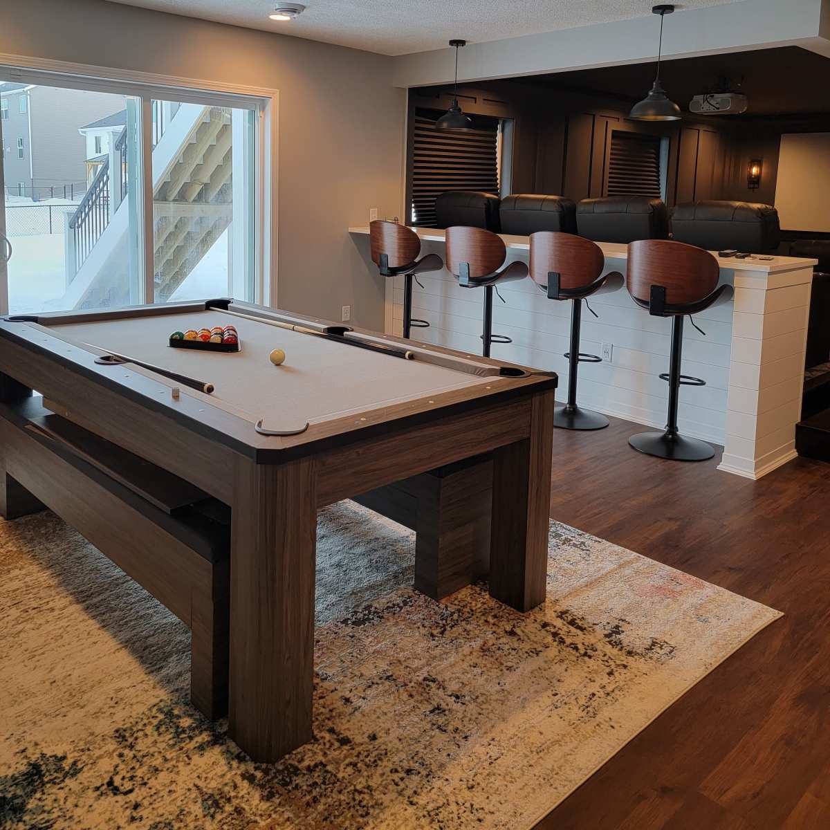 A modern game room featuring a pool table and a bar area with high stools, illuminated by recessed lighting.