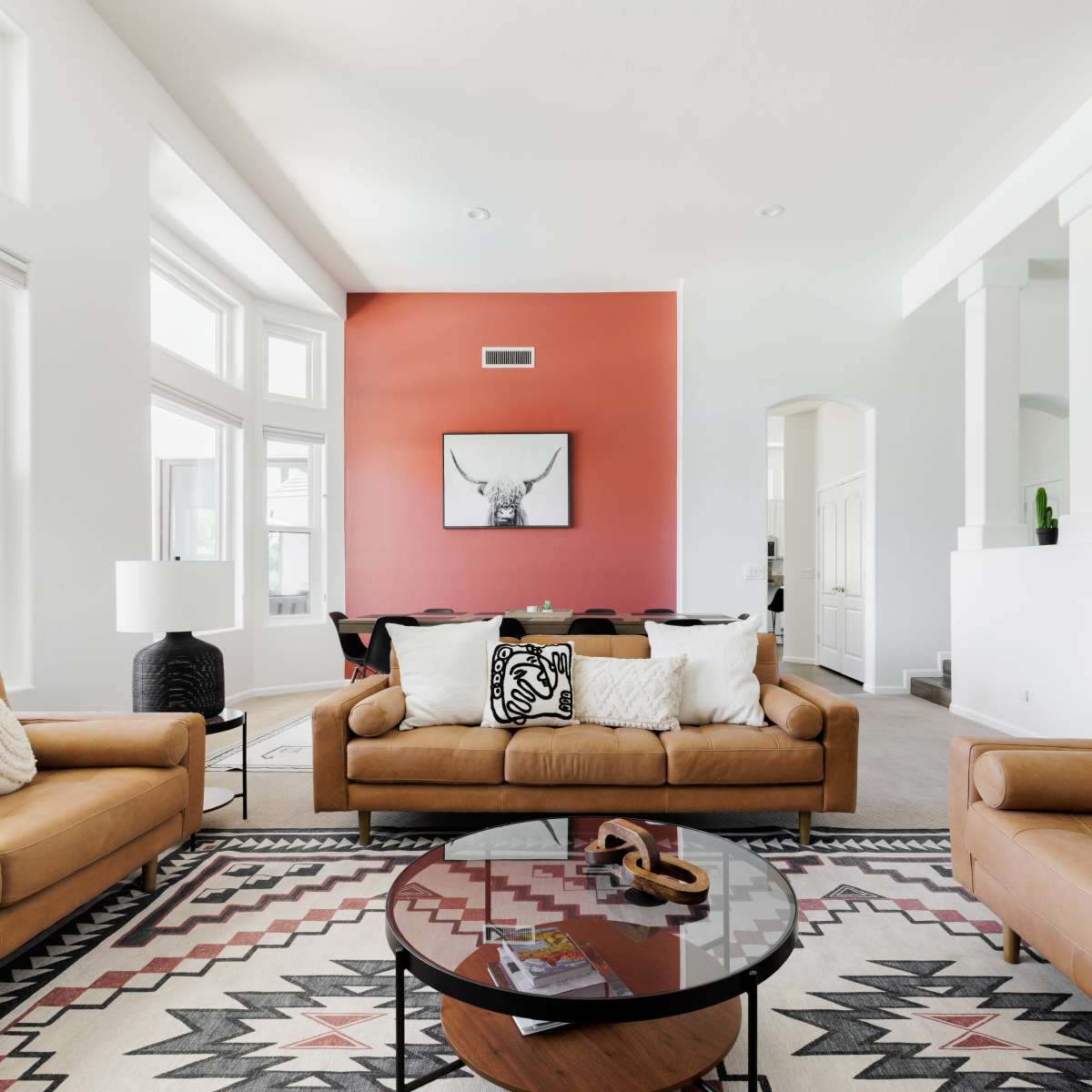 The living room features two brown leather sofas facing a round coffee table on a patterned rug, with large windows allowing natural light and a red accent wall displaying an artwork.