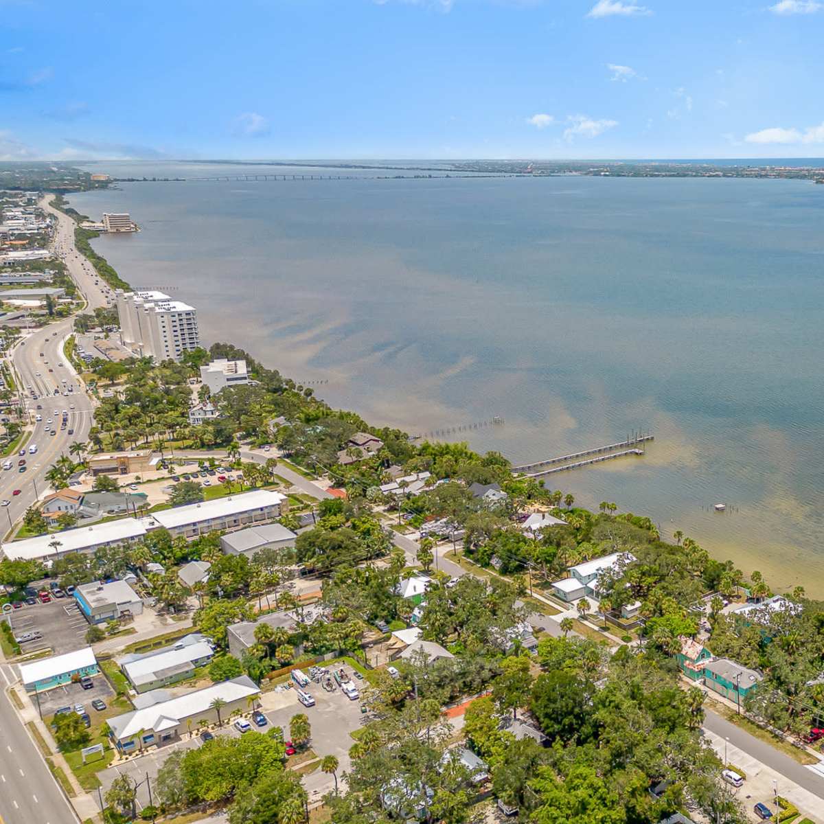 An aerial view captures a coastal area with nearby residential buildings, a dike extending into the water, and a city skyline in the distance.