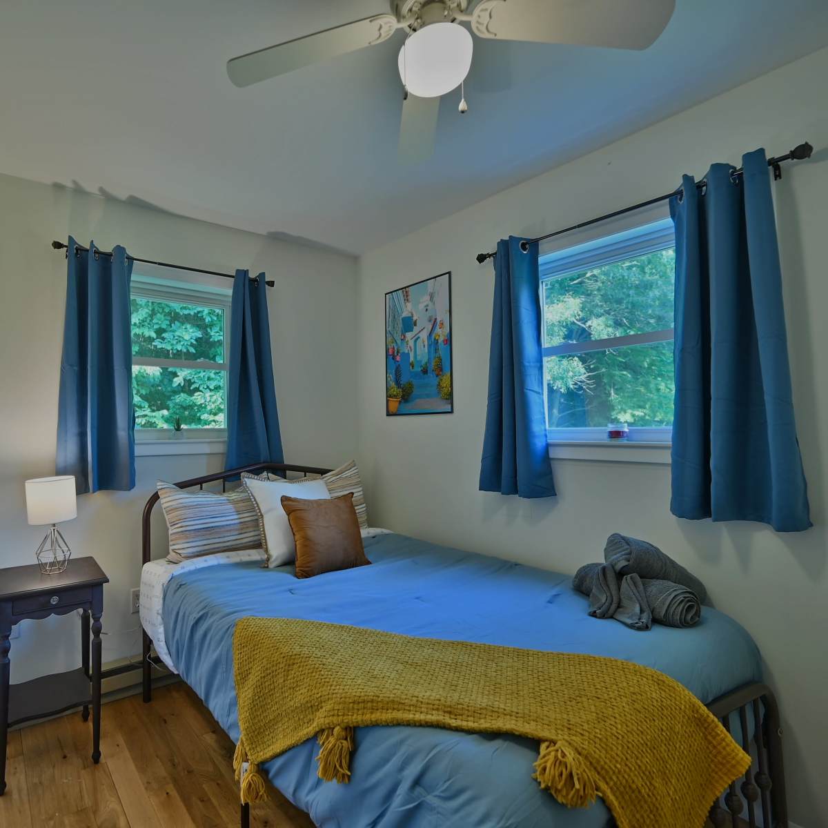 A small bedroom with a bed, bedside table, and two windows, adorned with blue curtains and decorative wall art.