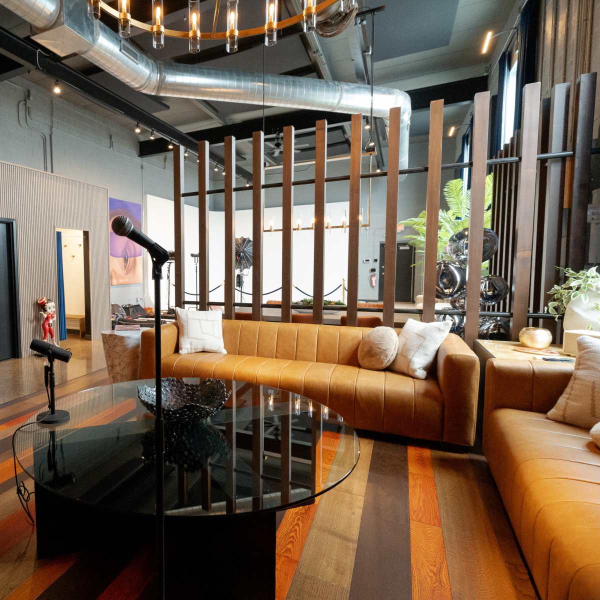 A modern lounge area featuring a circular glass table, a brown sectional sofa, and a microphone stand in the center, with a backdrop of wooden partitions and grey walls.