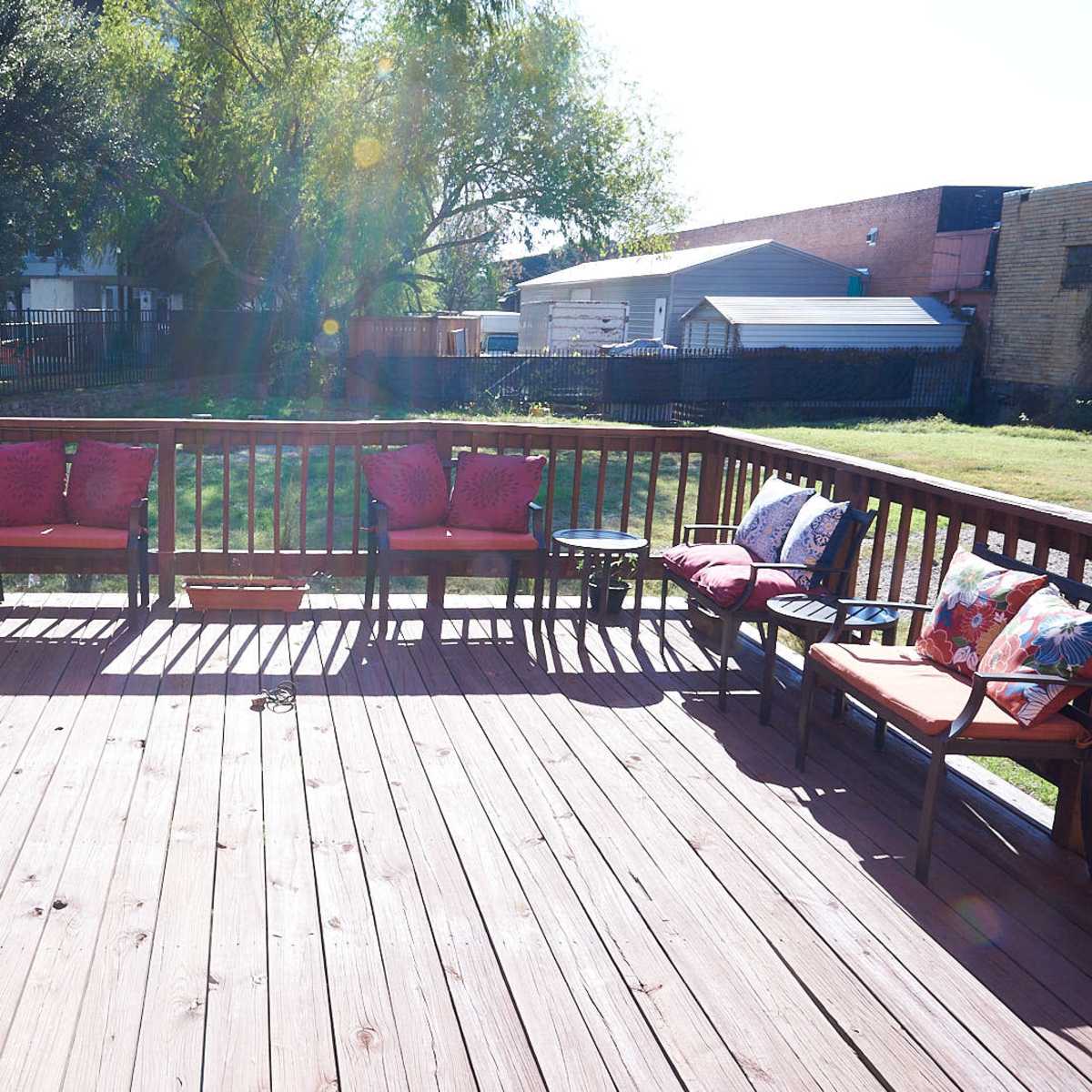 A wooden deck with several chairs and cushions arranged around the perimeter, surrounded by grass and trees.
