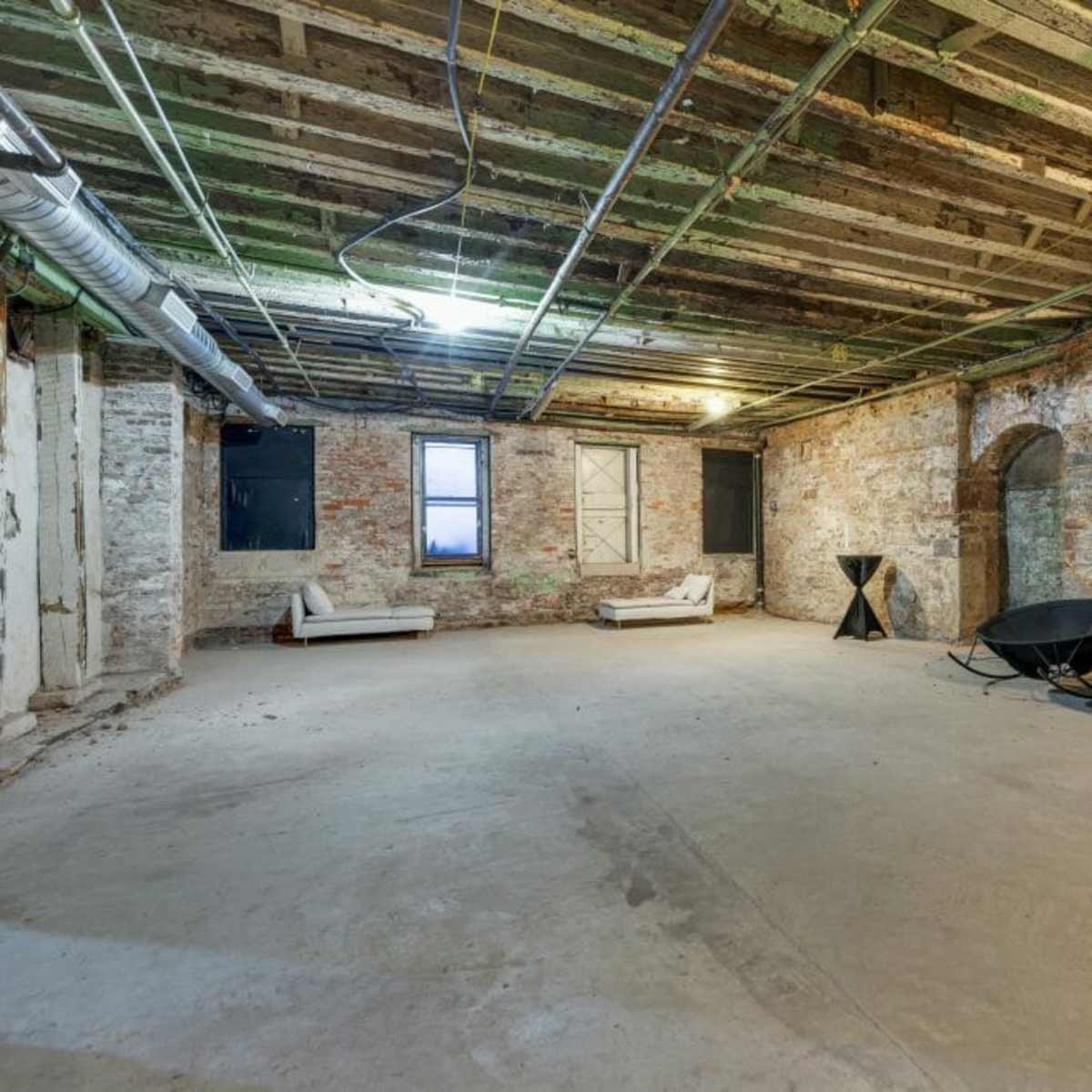 A spacious, unfinished basement features exposed brick walls, a concrete floor, and metal framework overhead.