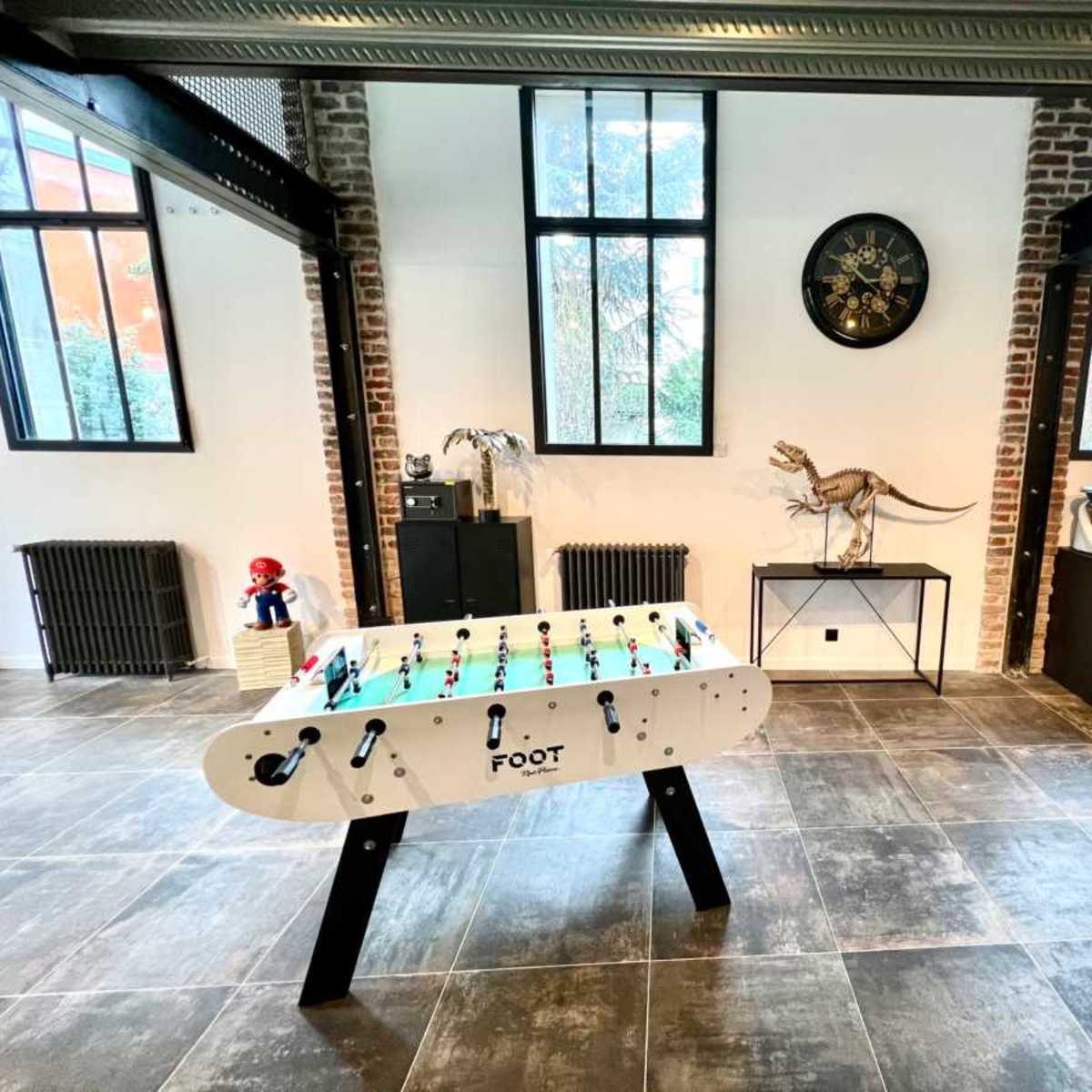 A spacious room with a foosball table in the center, large windows, and various decorative elements, including a dinosaur figurine and a modern sideboard.