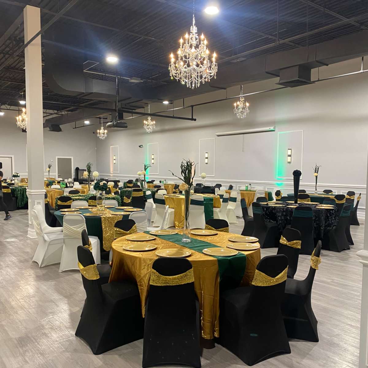 A banquet hall arranged for an event, featuring round tables with gold and green tablecloths, black chairs, and chandeliers overhead.