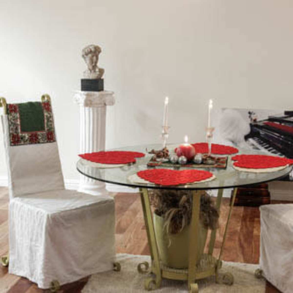 A circular glass dining table is set with red placemats and surrounded by two white-covered chairs, with candles and decorative items arranged in the center.