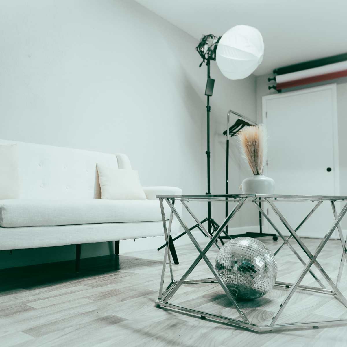 A light-colored sofa is positioned in a room with a reflective glass table that holds a decorative object, while a softbox light stands nearby.