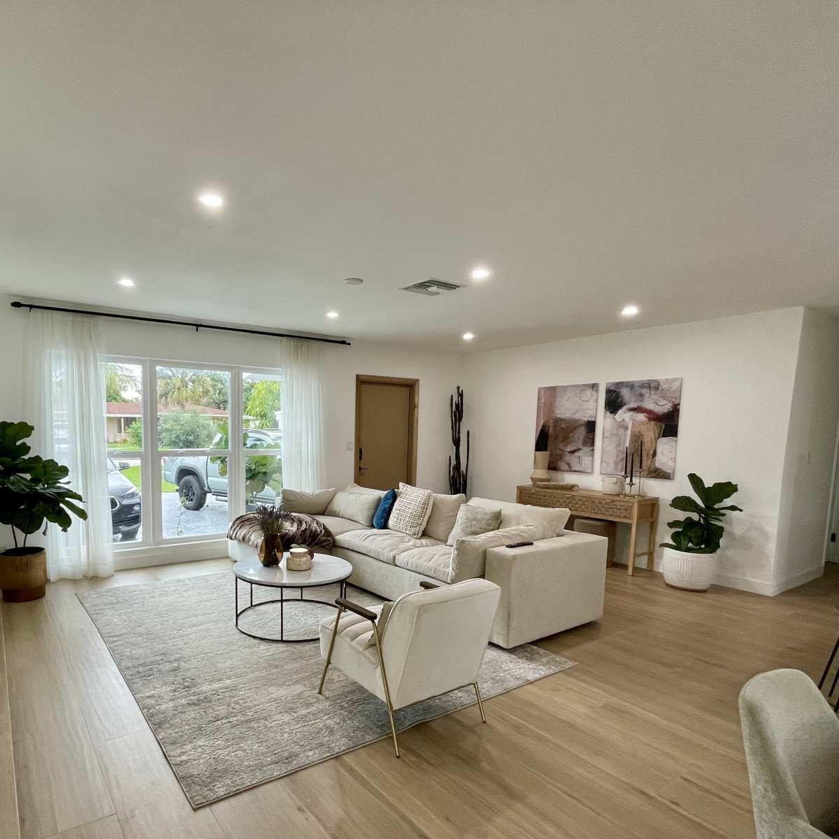 The living room features a light-colored sectional sofa, a coffee table, and large windows with sheer curtains, allowing natural light to illuminate the space.