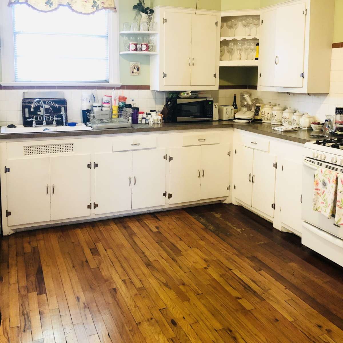 A kitchen with white cabinetry, a window adorned with floral curtains, and wood flooring.