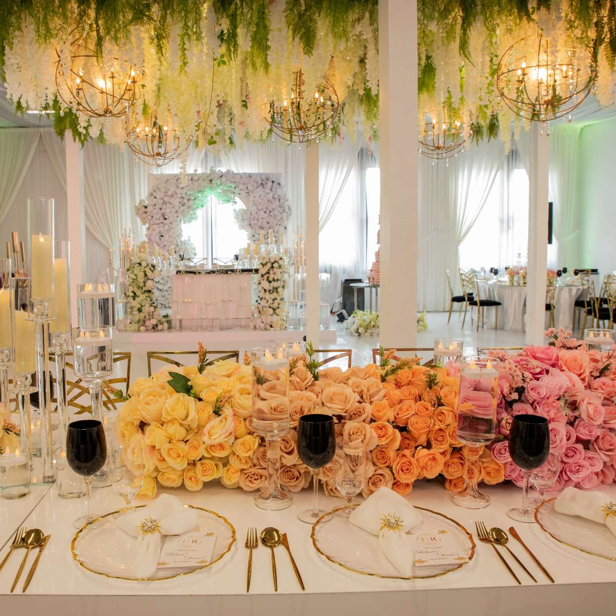A beautifully decorated indoor venue with an elaborately arranged table featuring pastel-colored floral centerpieces and elegant table settings under hanging greenery and chandeliers.