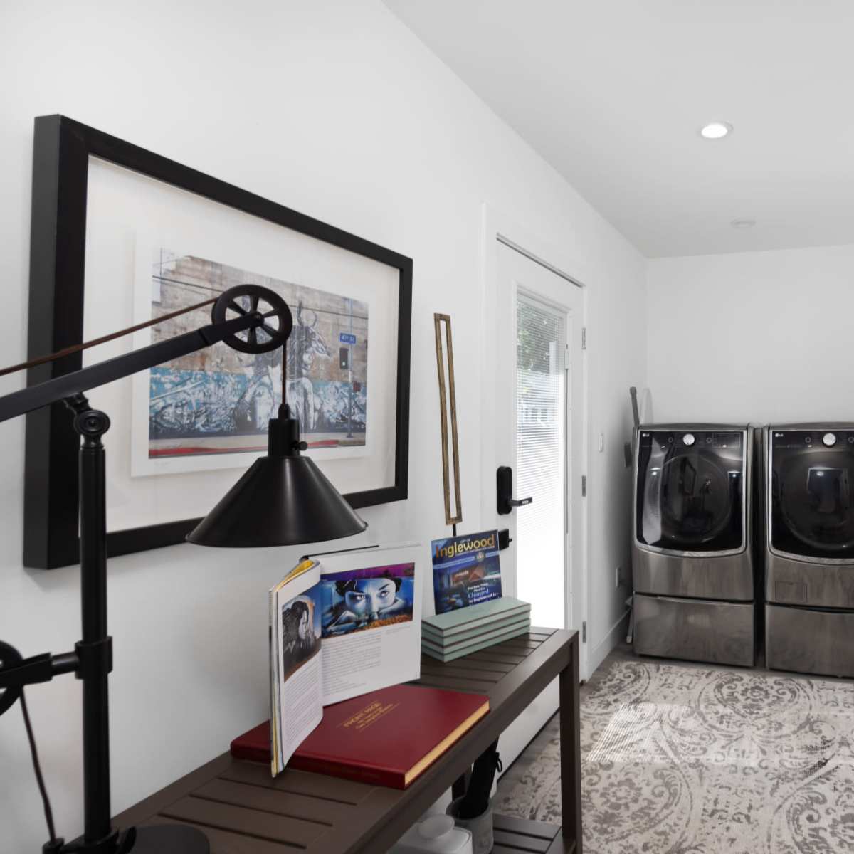 A laundry room featuring two black washing machines, a small table with books and a lamp, and a door leading outside.