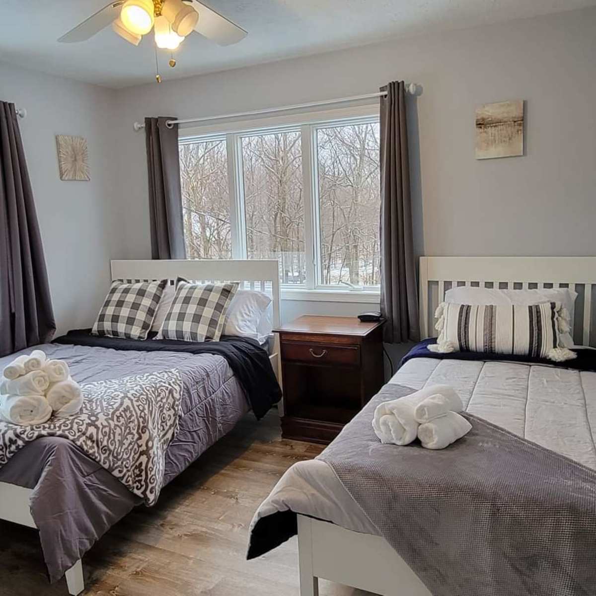 A well-lit bedroom with two beds, each neatly made with gray and white bedding, and a nightstand between them.