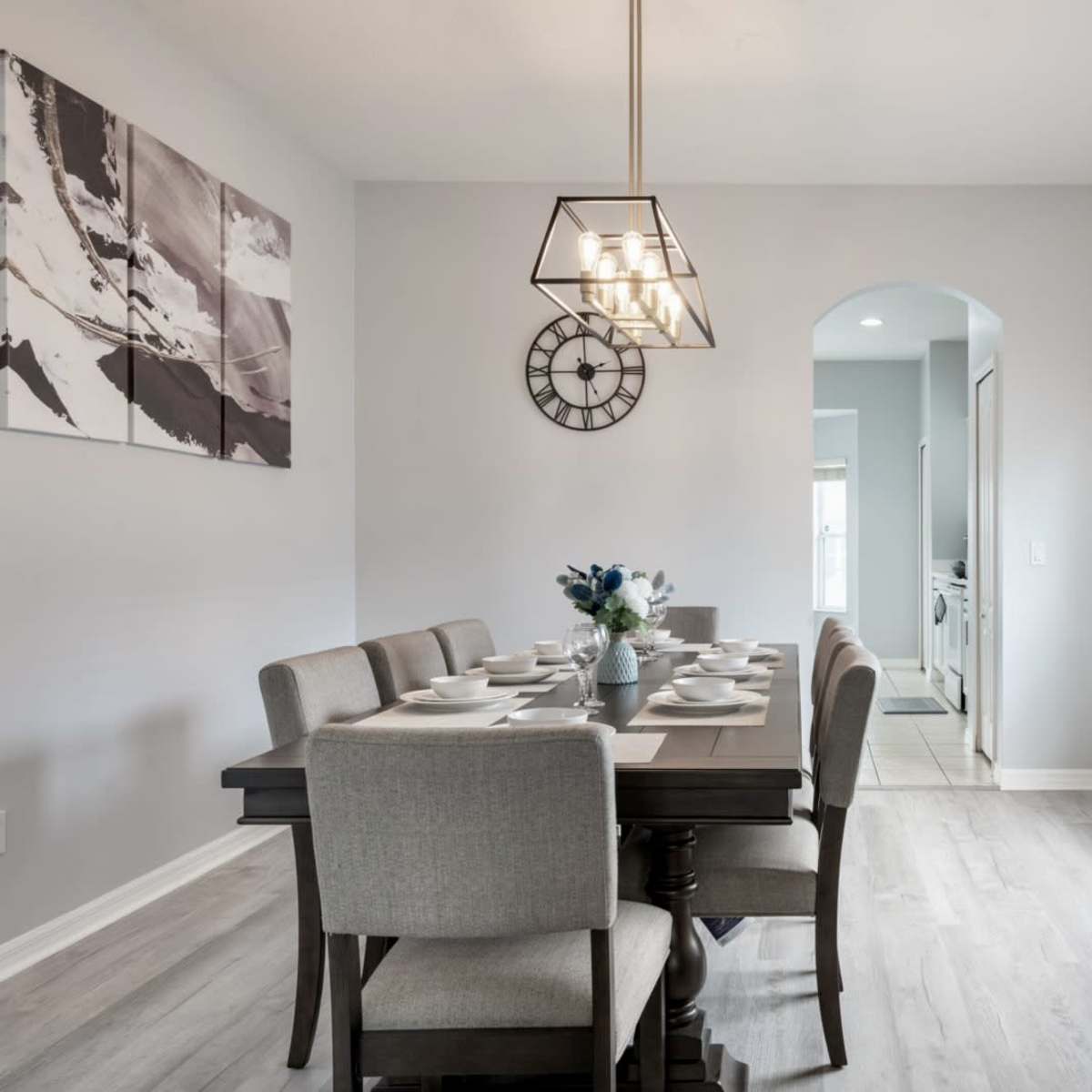 A dining area features a long table set for a meal, with a modern light fixture hanging above, and a clock on the wall beside an archway leading to another room.