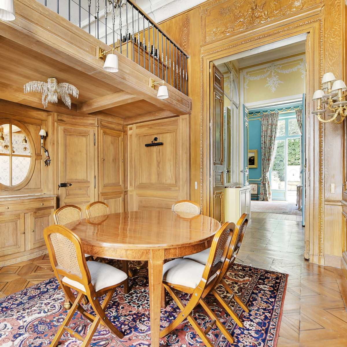 A wooden dining area featuring a round table surrounded by four chairs, with a staircase and decorative walls visible in the background.