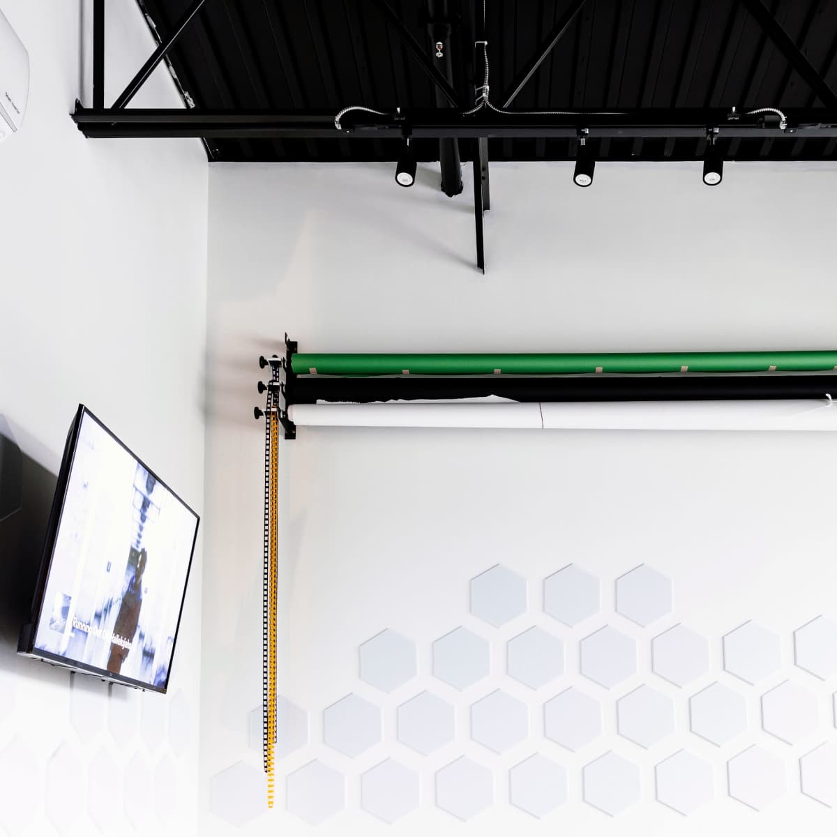 A white wall with hexagonal patterns, a mounted television, and a roll of green and white backdrop materials hanging from the ceiling.