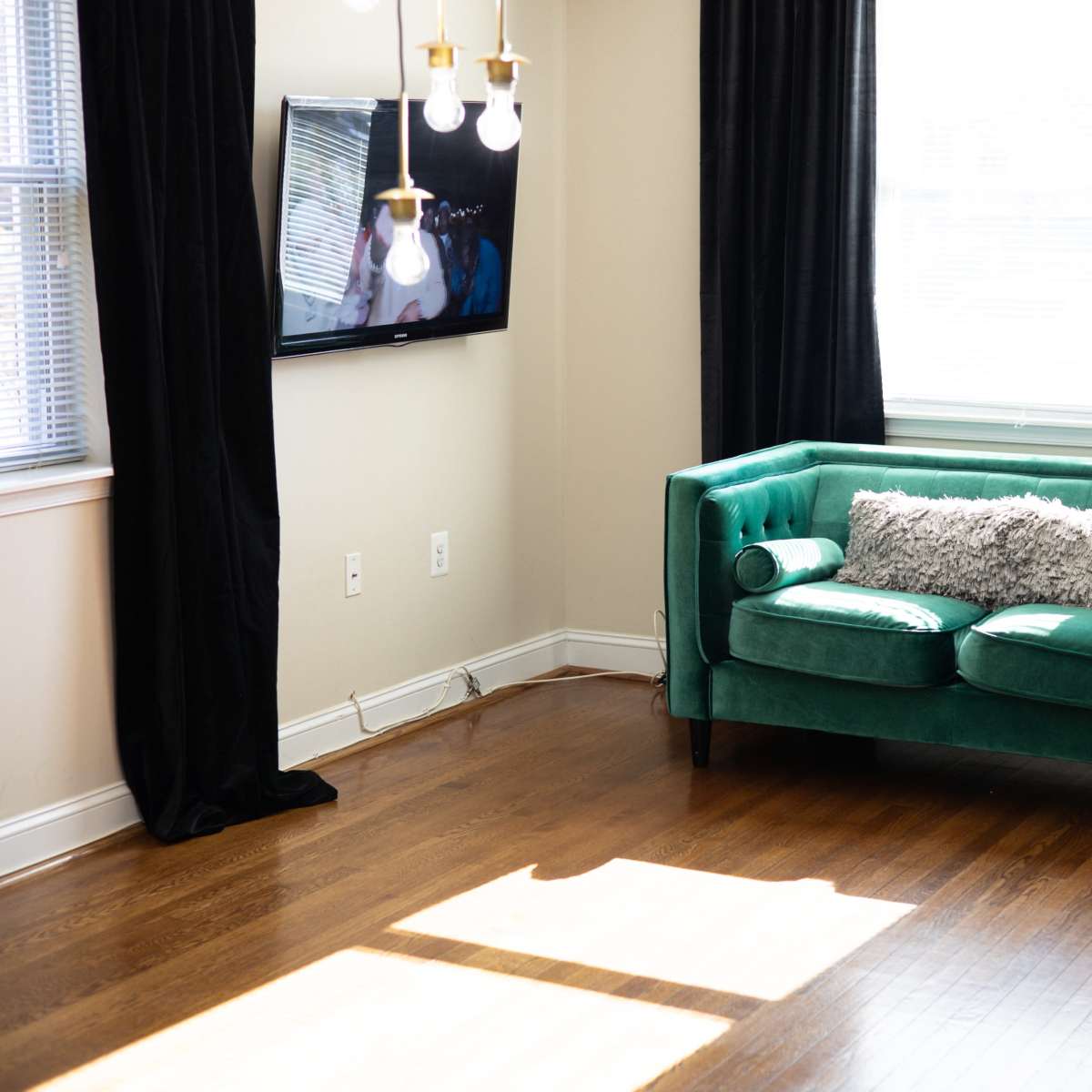 A green couch with decorative pillows is positioned in a sunlit corner of a room with hardwood floors and large windows.