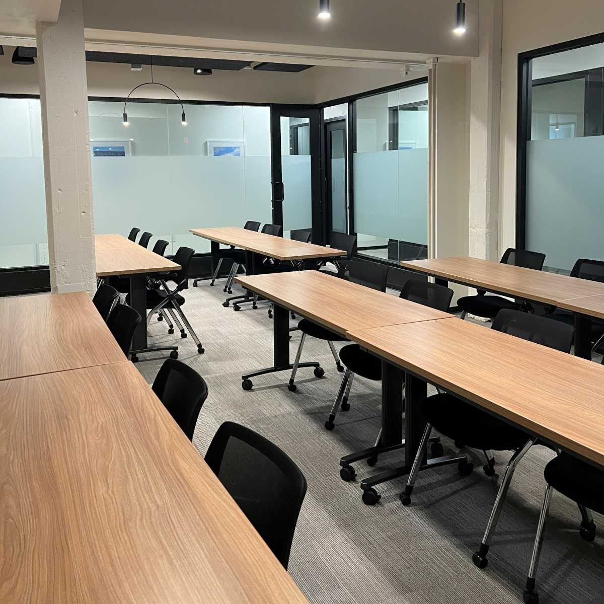 A modern conference room with several rows of long wooden tables and black rolling chairs, illuminated by overhead lights.
