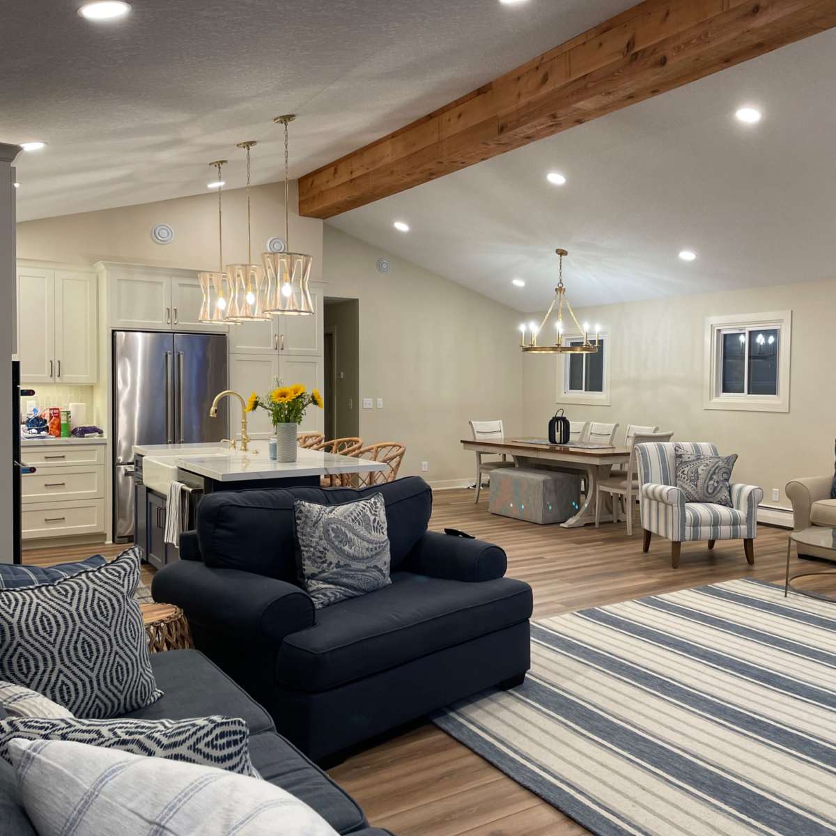 A modern living space featuring a combination of a kitchen and living area, with seating arrangements including sofas and a dining table under wooden beams and bright ceiling lights.