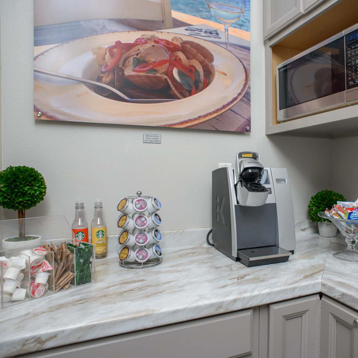 A small kitchen counter with a coffee maker, a microwave above, a bowl of snacks, and a display of drinks and utensils.