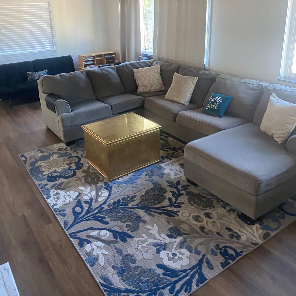 A spacious living room featuring a large gray sectional sofa, a gold coffee table, and a patterned area rug, with ample natural light coming through the windows.