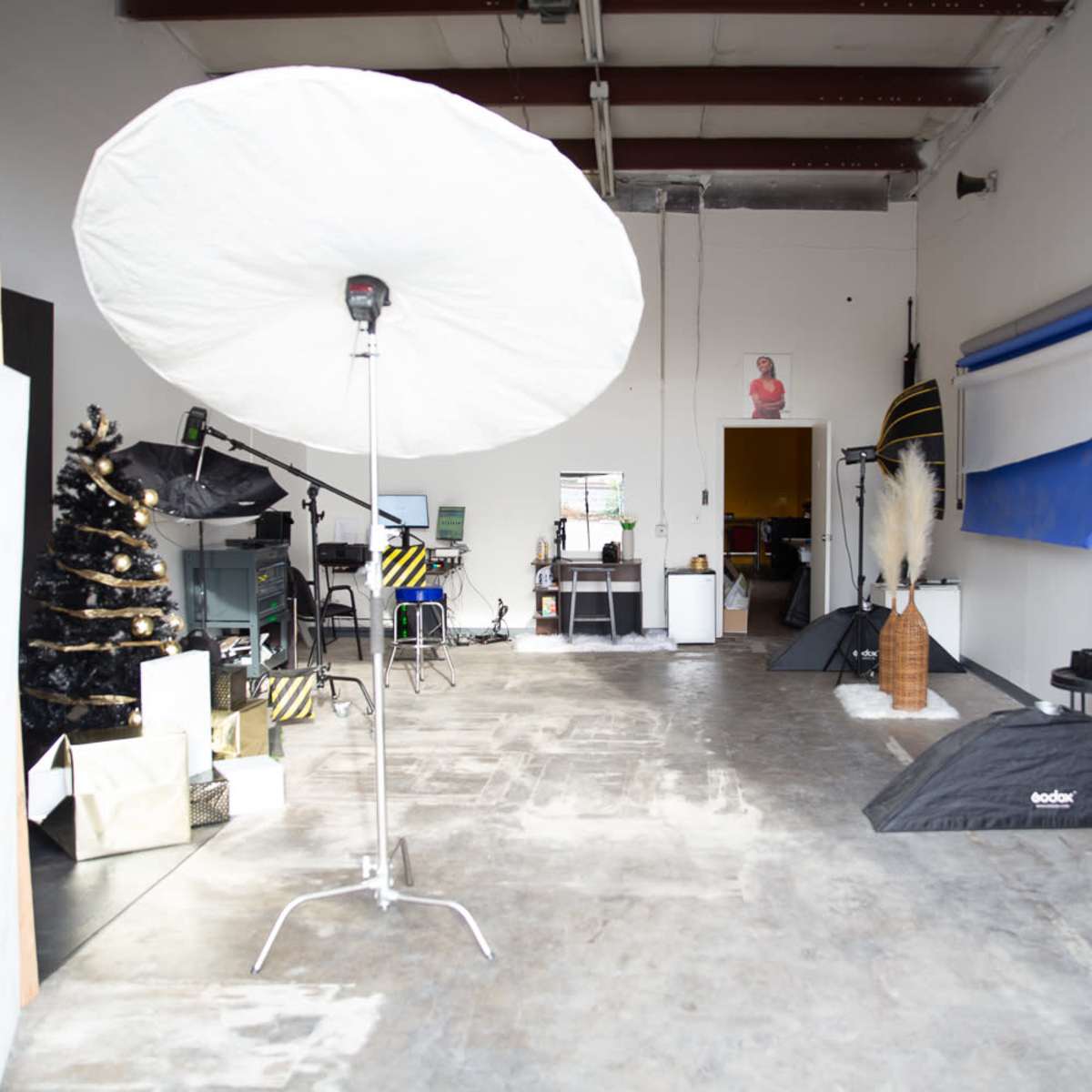 A photography studio featuring a large umbrella light, a black Christmas tree, and various props and backdrops arranged throughout the space.