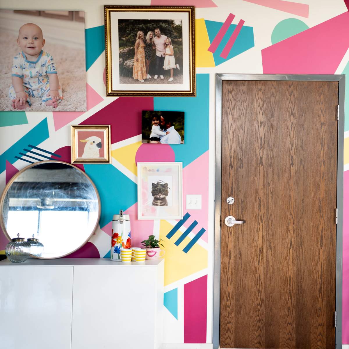 A vibrant wall with a geometric design in pink, turquoise, and yellow, adorned with framed photos and a round mirror, alongside a wooden door and a shelf displaying various decorative items.