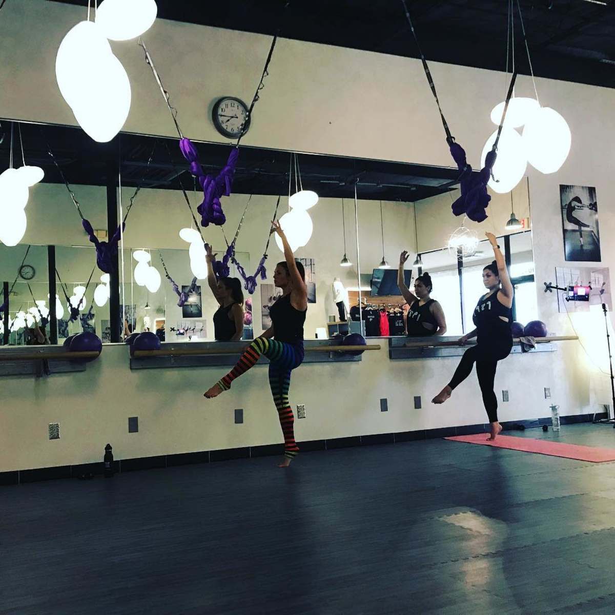 A group of four women performing aerial yoga in a studio with purple hammocks and mirrored walls.