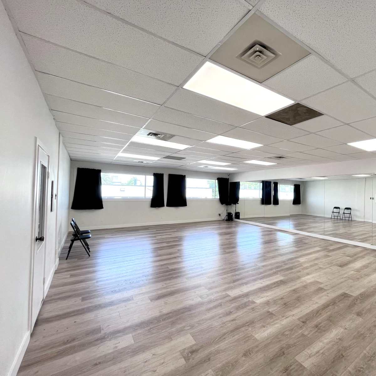 A spacious, empty room with wood-like flooring, large windows covered by black curtains, and a full-length mirror on one wall.