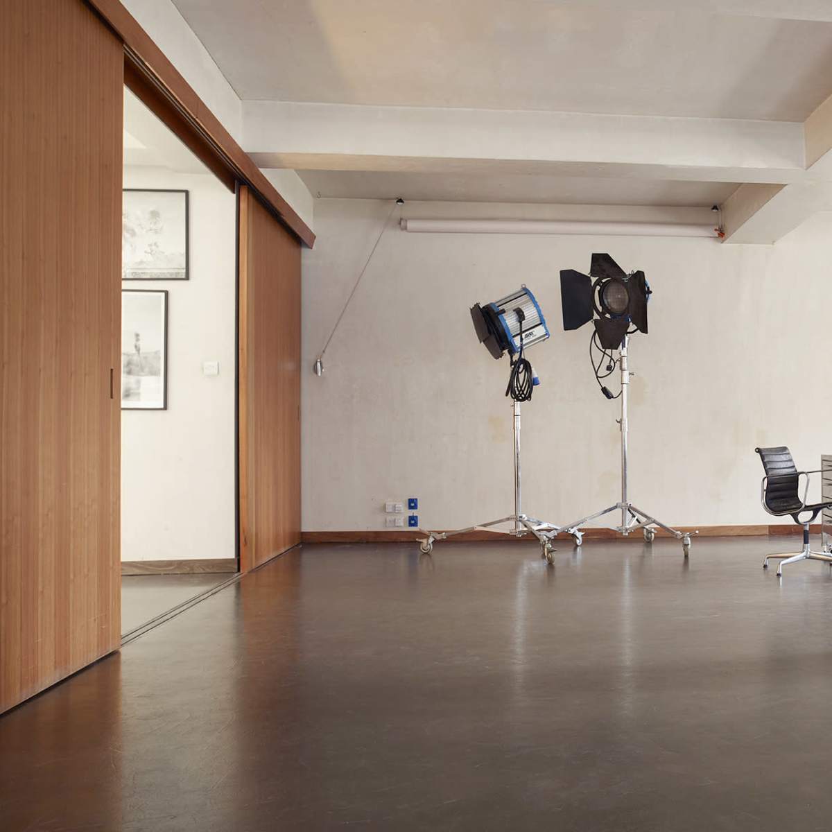 A spacious, minimalistic studio with a desk, a chair, and several lighting equipment positioned in the corner.