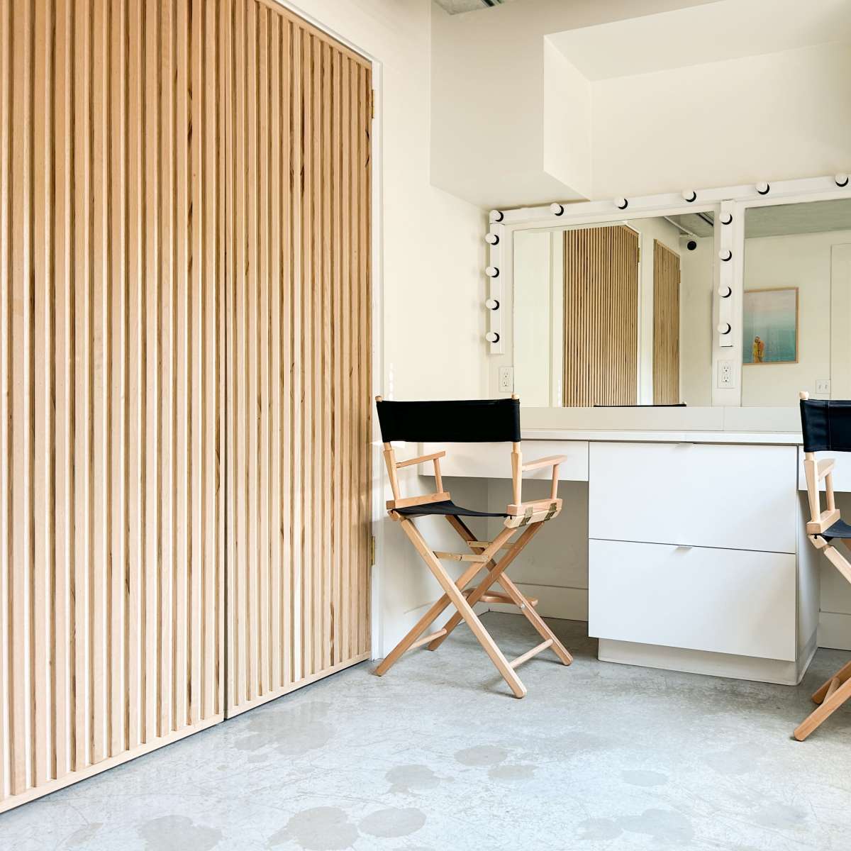 A minimalistic dressing area with two director's chairs and a large mirror surrounded by lights, set against a wooden door.
