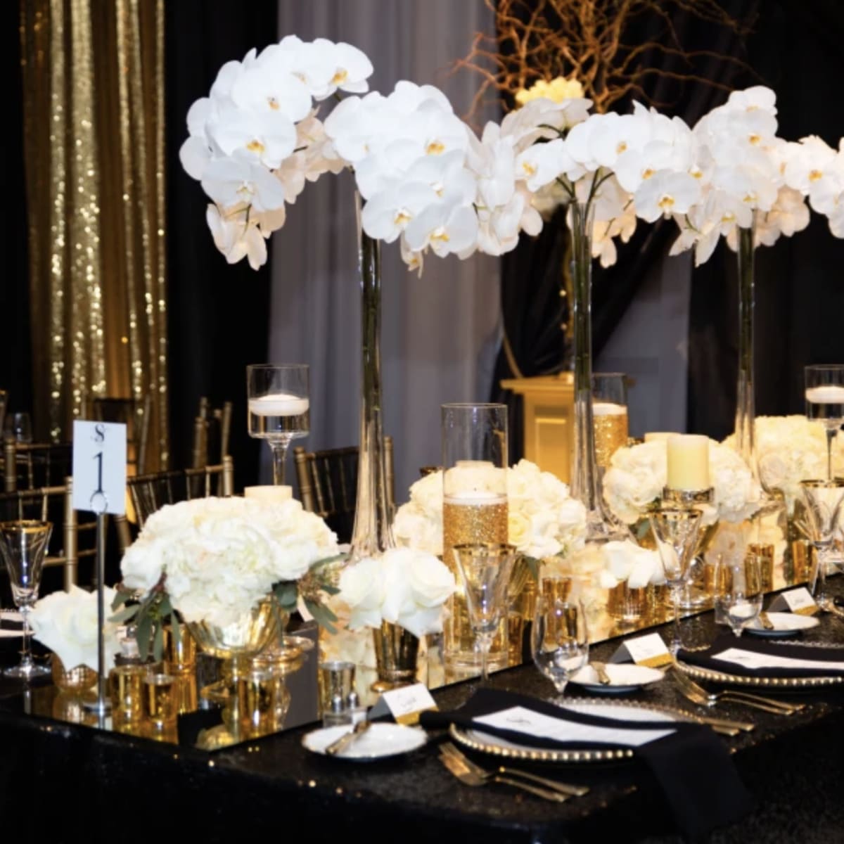 A elegantly set dining table features tall white orchid centerpieces, gold accents, and crystal glassware against a backdrop of black and gold decor.
