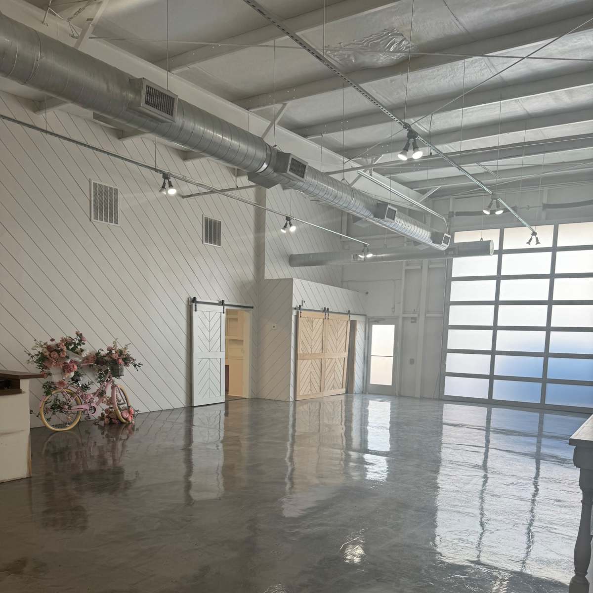 A wide, empty room with polished concrete floors, white walls, and large windows, featuring a pink bicycle and floral decorations in one corner.