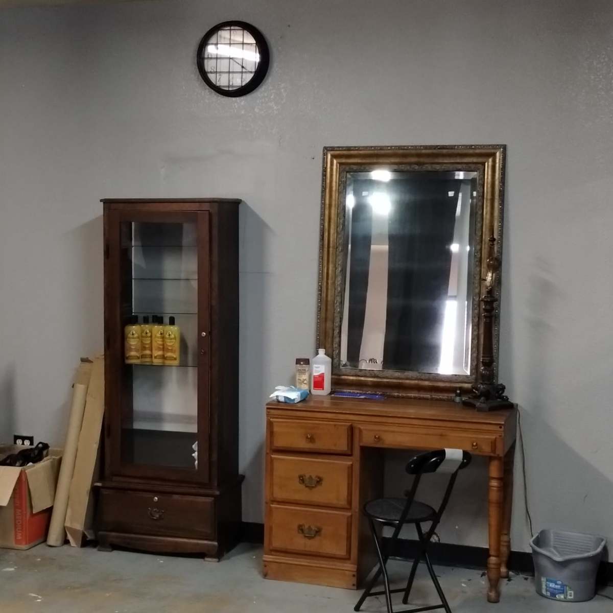 A garage space with a gray wall, a circular window, a glass-front cabinet, a wooden vanity, a white chair, and various items along the floor.